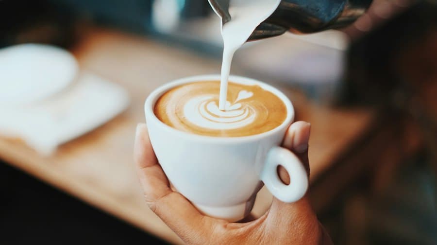 A cup of latte with milk being poured into it, held in one hand