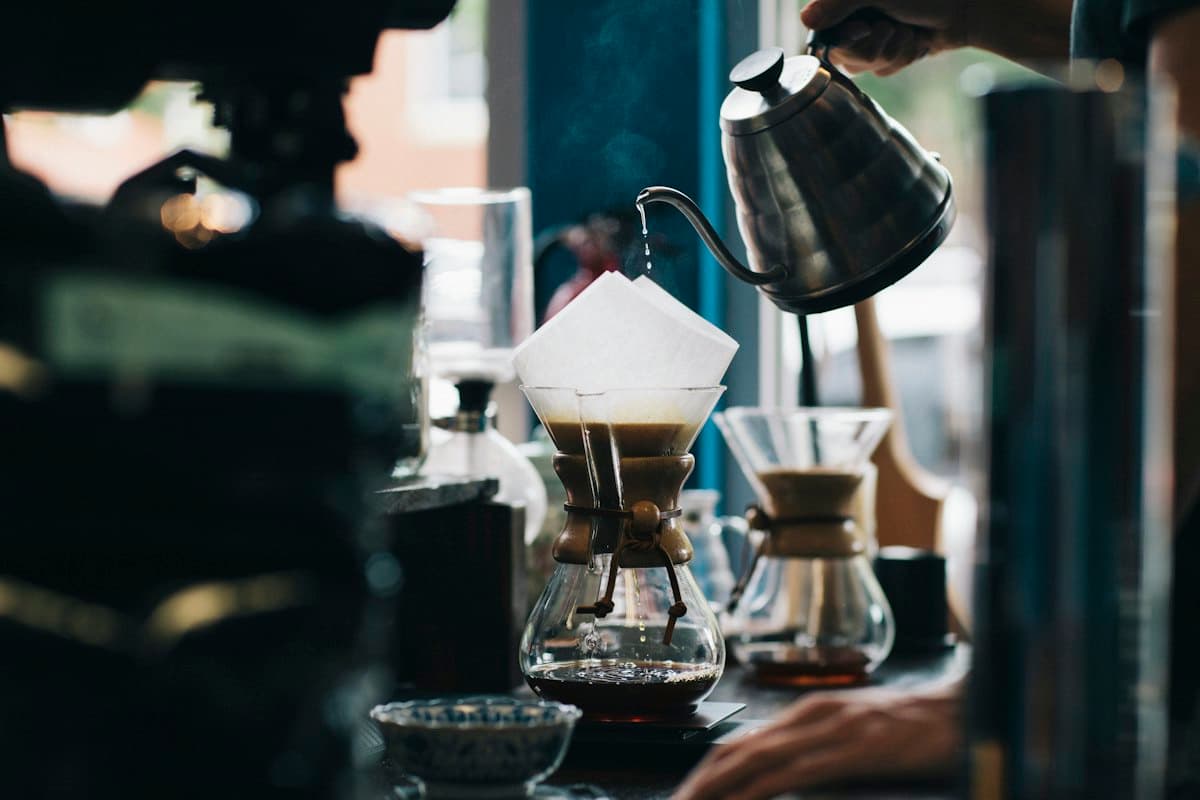 A barista pouring hot water over a pour-over coffee setup by the window