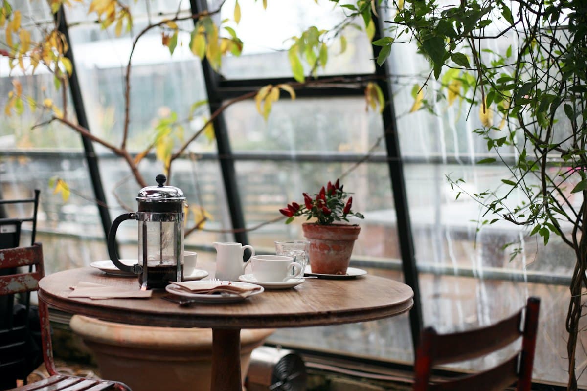 A round wooden table by a sunlit window with a french press and coffee cups
